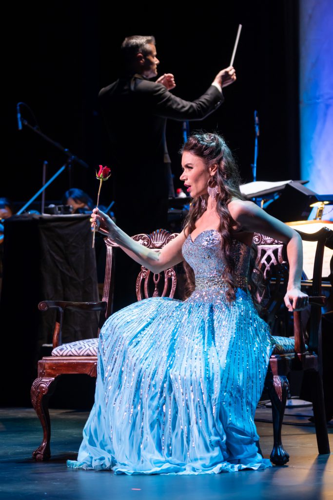 Singer Ginger Costa-Jackson holds a rose while she sings in a light blue sparkling gown, with conductor David Charles Abell conducting behind her. 