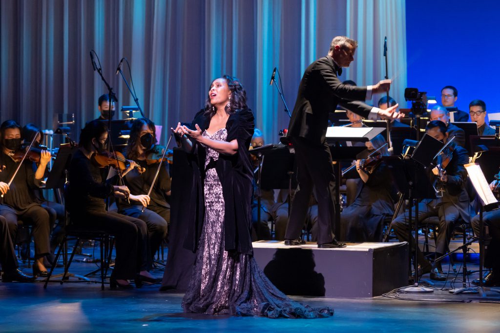 Singer Talise Trevigne, in a black velvet cloak and black lace gown, performs on stage with the Kansas City Symphony, conducted by David Charles Abell. 