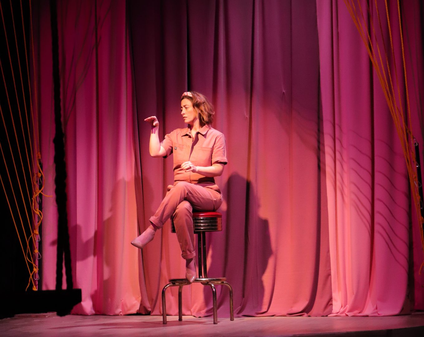 A woman sits on a stool on a stage, talking to her own hand.