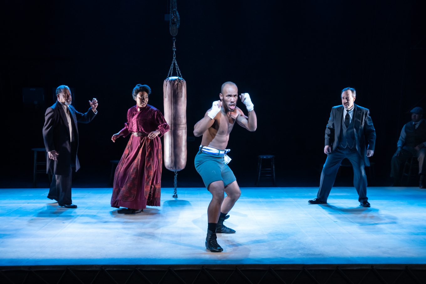 Dressed in 1910s costumes, a Black boxer stands in fight stance center stage, surrounded by other characters in The Royale