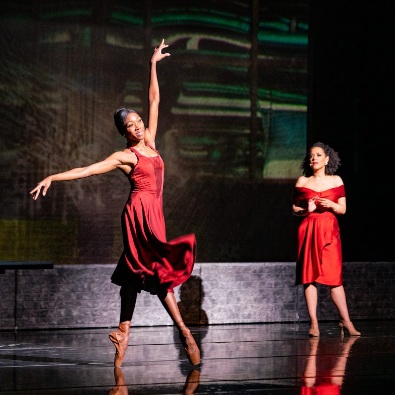 Dancer in a red dress en point with woman in a red dress watching her.