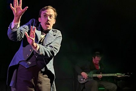 An actor gestures while another man plays sits and guitar in the background.