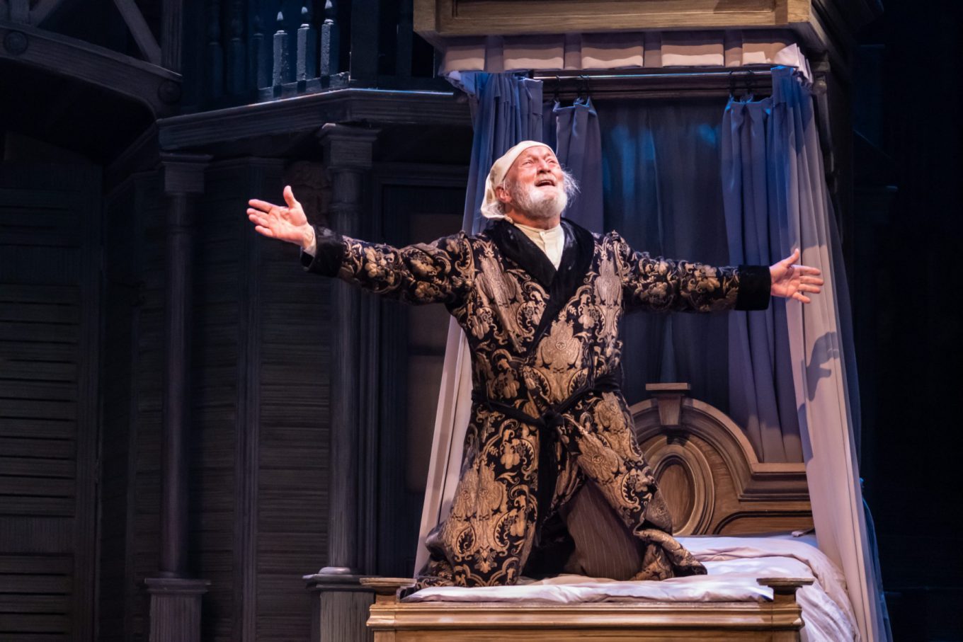 An older white man in an old-fashioned dressing gown and night cap kneels on a bed with his arms outstretched, looking up and grinning.