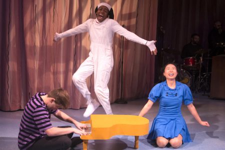 An actor dresed as Snoopy dances on a small yellow piano while another actor plays it and another holds her arms out in joy, kneeling on the ground.