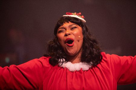 A young Black woman in a red Christmas outfit belts while singing.