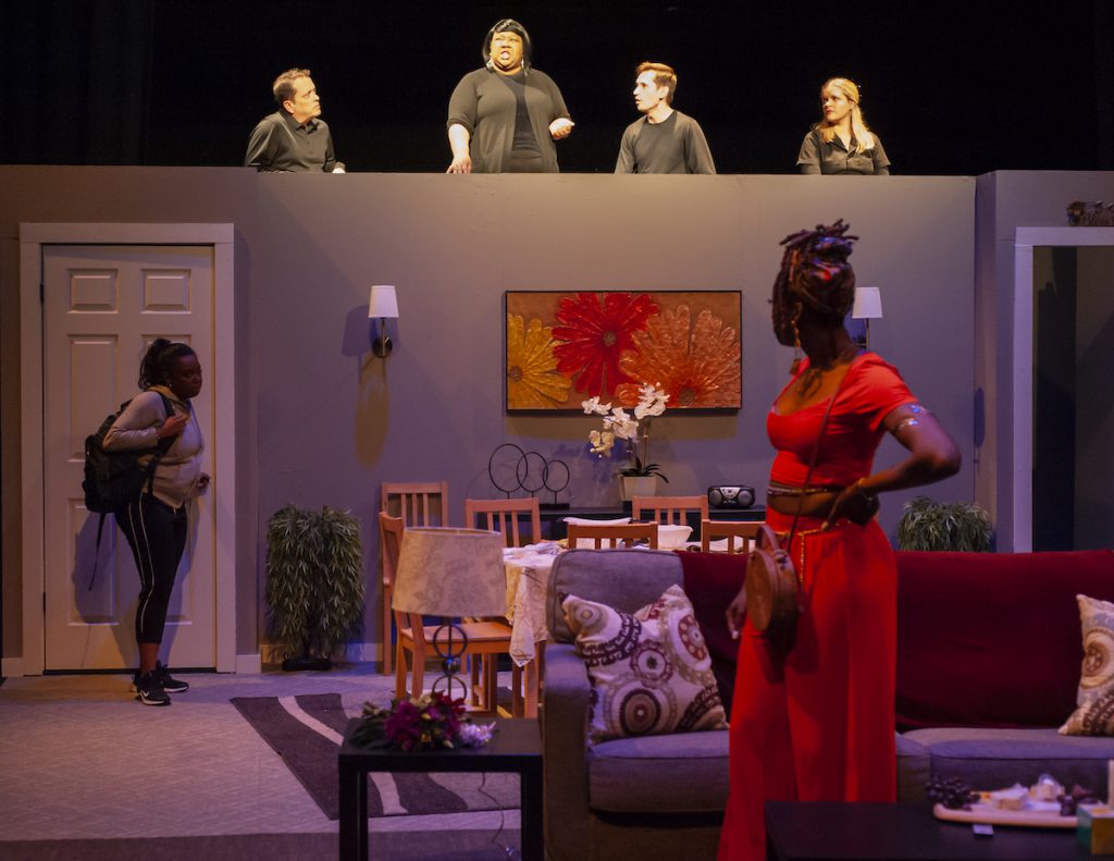 Two Black actors onstage act out a scene while a group of other actors dressed in black observes from above.