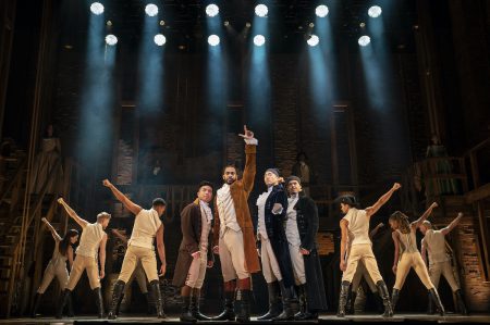 The cast of Hamilton faces forward, raising hands into the air triumphantly.