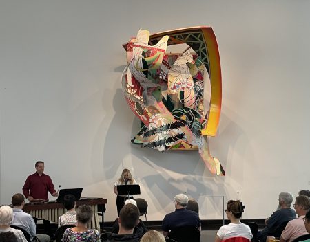 Kevin Clarke performs on marimba and Sharra Wagner performs on clarinet in front of a large multicolored sculpture by Frank Stella.