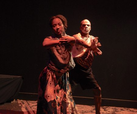 A Black woman dances as a Black man stands behind her, mirroring her movements.