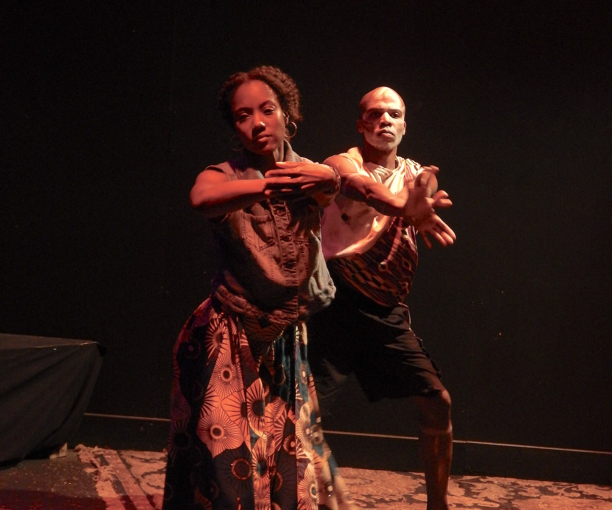 A Black woman dances as a Black man stands behind her, mirroring her movements.
