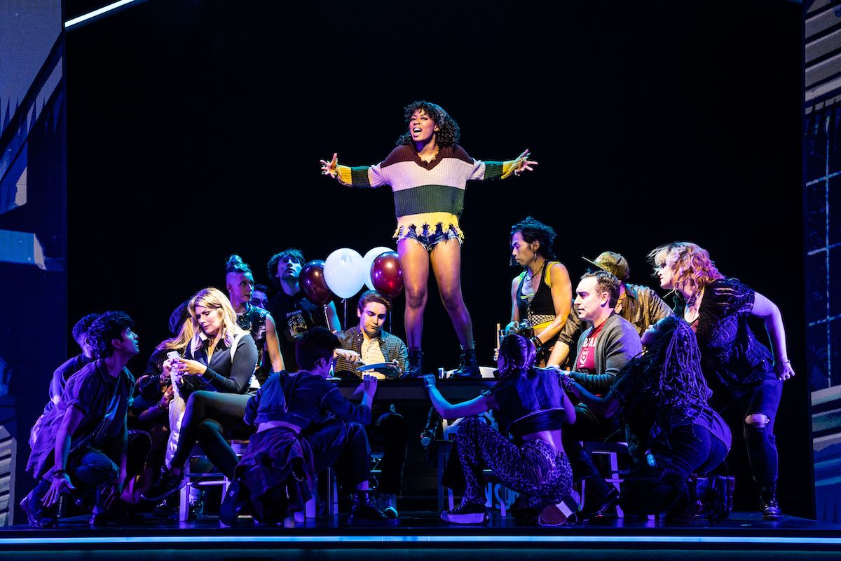 In a scene from Jagged Little Pill, a young Black woman (Frankie) stands on a table with her arms outstretched, surrounded by a crowd of other characters.