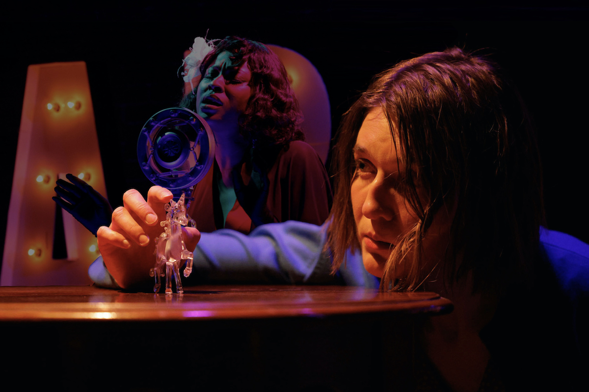 A young woman leans her chin on a table, looking at a glass unicorn. A beautiful Black woman can be seen behind her singing jazz