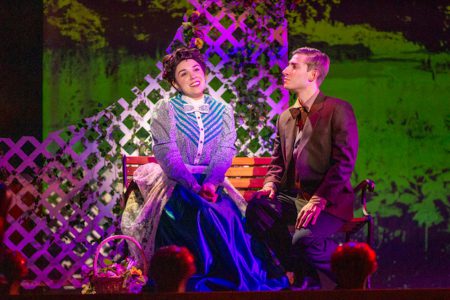 In a scee from a play, a young man and woman dressed in early 20th Century costumes perform a romantic scene in front of a trellis.