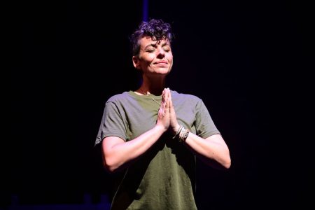 A person gives a strained smile, eyes closed, with hands in front of them in prayer position.