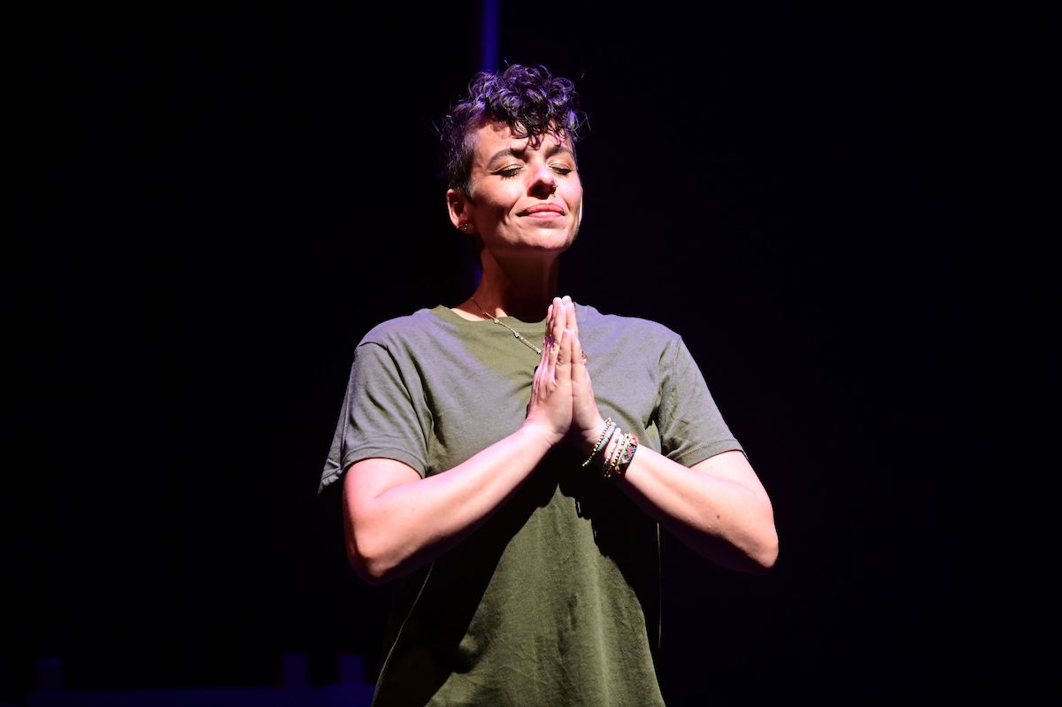 A person gives a strained smile, eyes closed, with hands in front of them in prayer position.