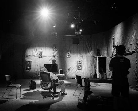 A black and white photo of a room with speakers all along the walls. Two figures study the space.