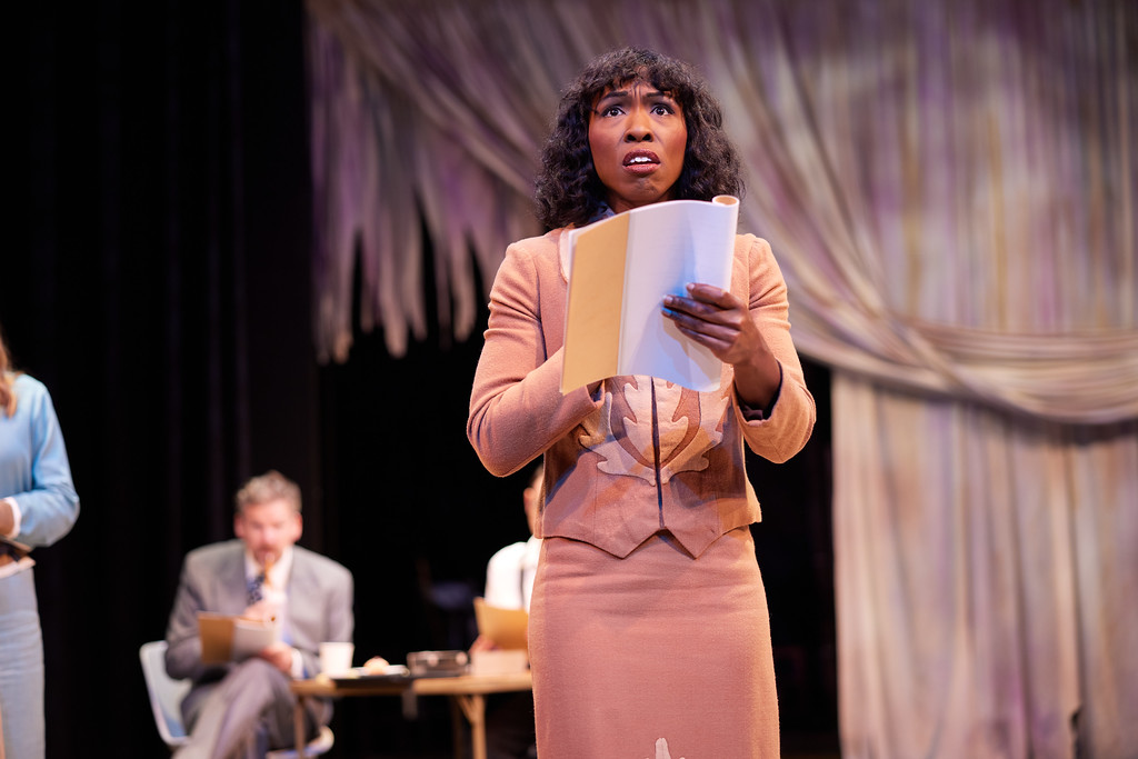 A Black woman dress in 1950s clothing, reading a script dramatically