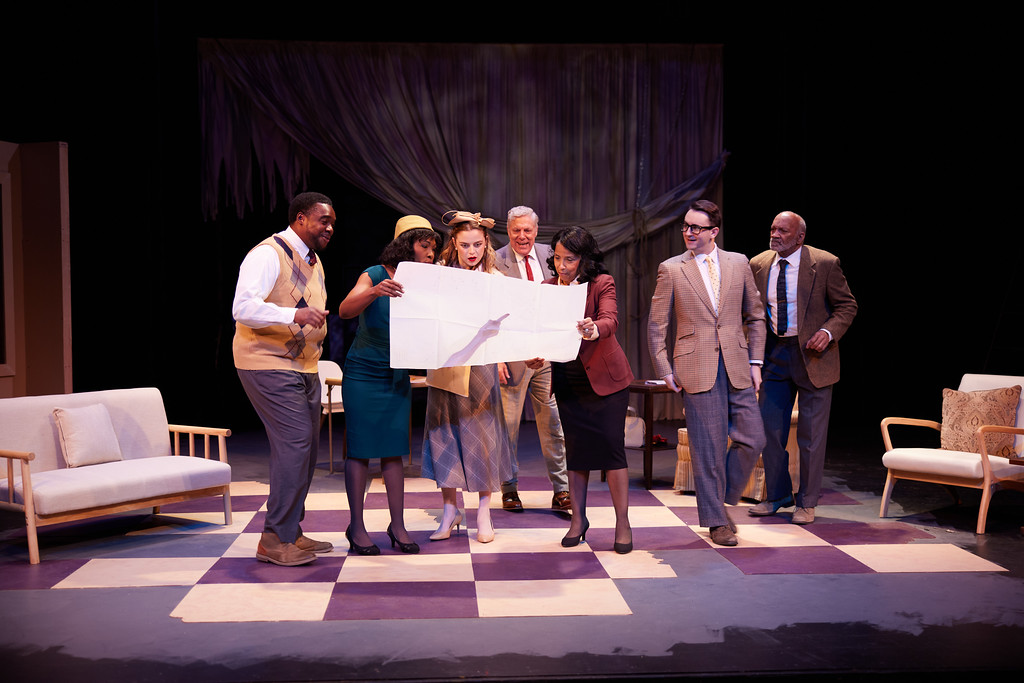 A group of actors playing 1950s stage actors gather to look at blueprints.