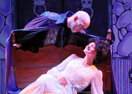An actor playing Dracula leans over a woman in 19th century clothing reclining on a bench.