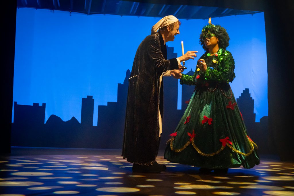 An actor playing Ebenezer Scrooge and an actress as the Ghost of Christmas Present, in a long dress resembling a Christmas Tree, talk animatedly on stage.