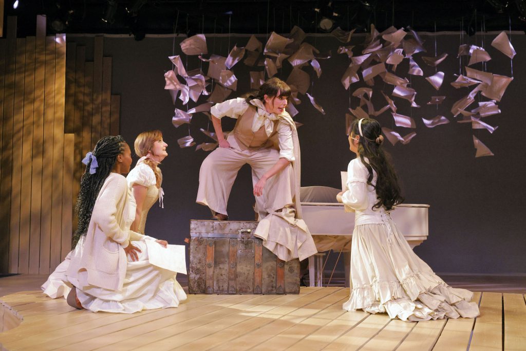 In a scene from Little Women, the sisters gather around Jo, standing on a trunk, commanding their attention.