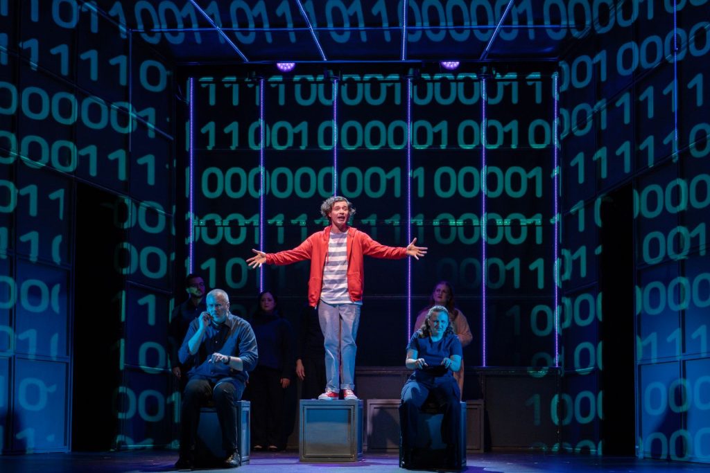 An actor stands on a block speaking animatedly. Ones and zeros are projected onto the walls behind him.