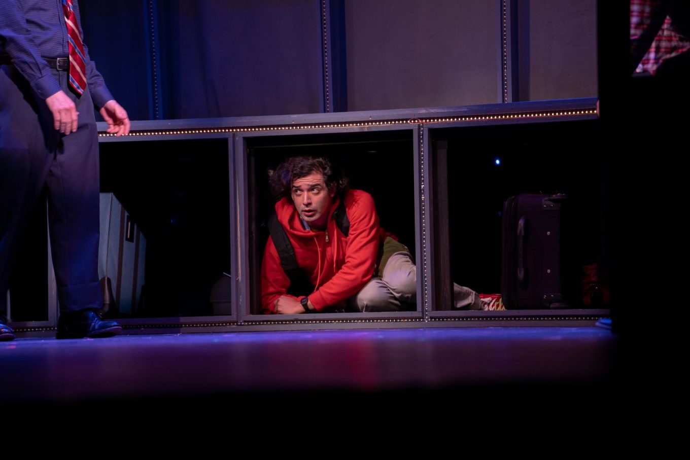 A young man in a red hoodie and a backpack hides in a train's luggage compartment.