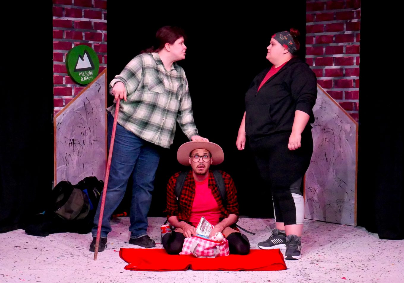Two actresses argue standing over a man sitting on a picnic blanket looking uncomfortable.
