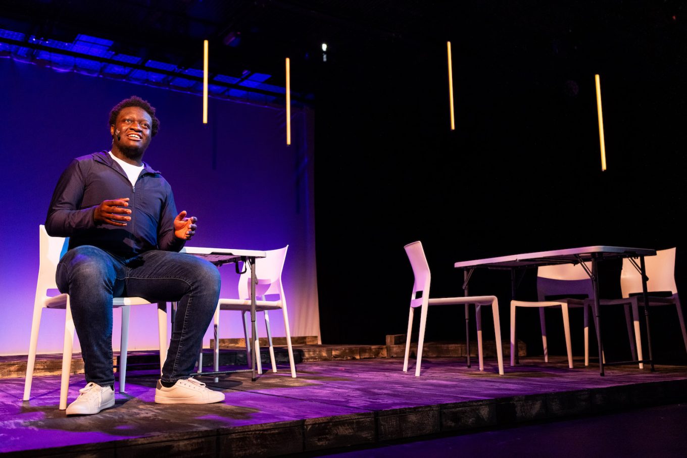 A Black teen sits onstage delivering a monolouge directly to the audience.
