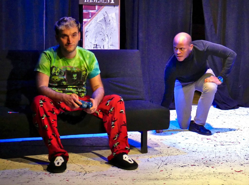 An actor plays video games while another crouches behind him.