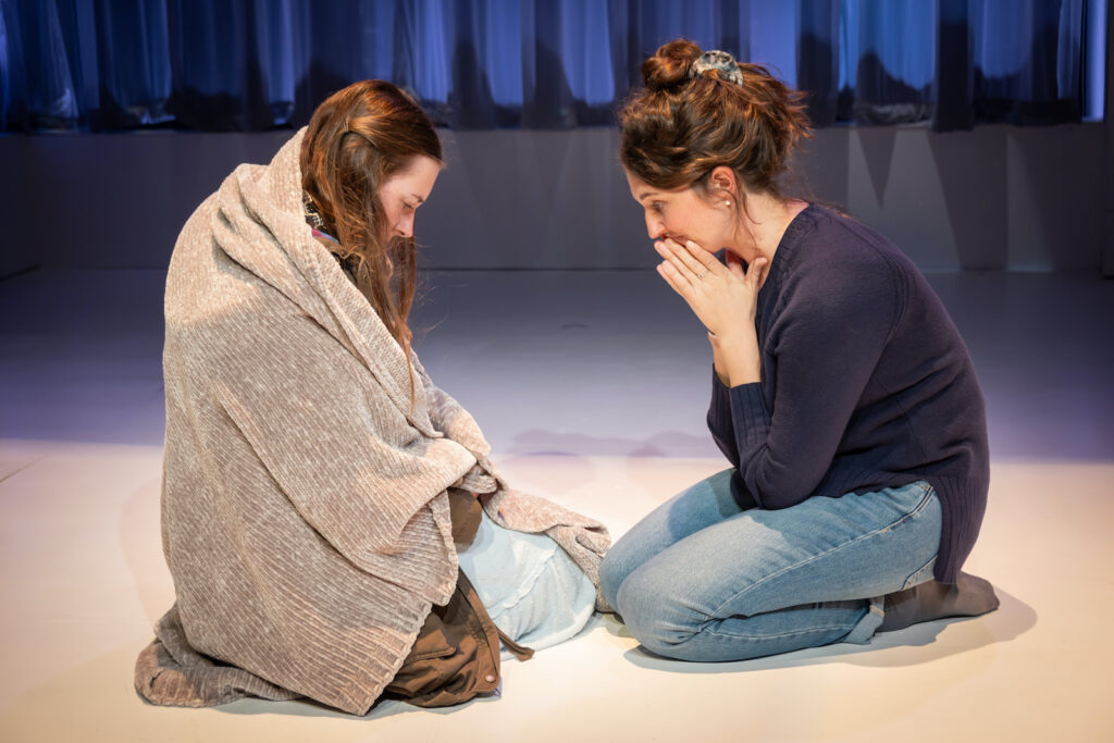 A young woman and a teen girl sit on the floor facing each other, looking down, both looking pensive.