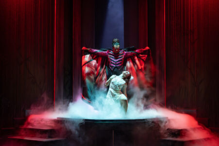 A woman kneels in front of Dracula, baring her neck to him. His arms are outstretched, holding out his cape. They're cast in red light as smoke pours out from between them.