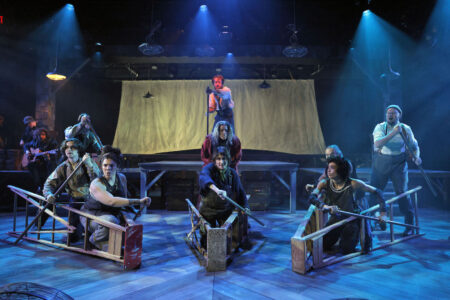 A group of actors composed in a tableau. Ladders are laying flat to resemble boats, with the actors sitting inside their openings.
