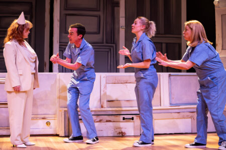 Three actors in matching blue jumpsuits gesticulate emphatically toward a woman in a white suit and a paper party hat.
