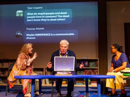 Three adults sit in front of a laptop in a children's school library with a livestream chat projected on the wall behind them.