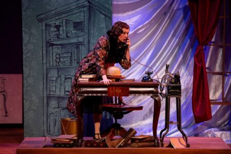 An actress playing Agatha Christie stands at an old timey writing desk, talking emphatically into a telephone.