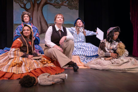 A group of actors in a scene from a play within a play of Little Women, sitting on the floor looking wistful.