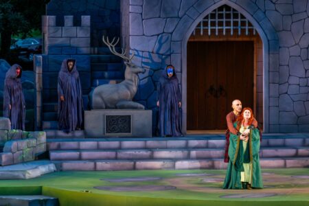 Macbeth and Lady Macbeth embrace downstage in front of a large castle set, while the witches look on from the shadows behind.
